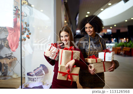 Two mixed race women with gift boxes in hands near storewindow. Multi ethnic girls smiling with presents on christmas new year sale. Caucasian and african american females buy presents for holidays. Two mixed race women with gift boxes in hands near storewindow. Multi ethnic girls smiling with presents on christmas new year sale. Caucasian and african american females buy presents for holidays. 113126568