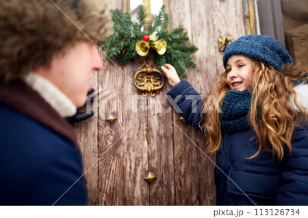 Little girl helps father and hanging Christmas wreath on the door. Cute curly daughter spend time with parents on holidays. New Year home decorations and preparations. Snowy winter outdoors. 113126734