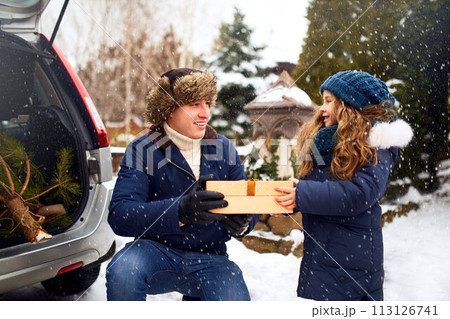 Father presents daughter a gift box on snowy winter day outdoors. Christmas tree in large trunk of family car. Girl helps dad with New Year home decorations and preparations for holidays. Snowfall. Father presents daughter a gift box on snowy winter day outdoors. Christmas tree in large trunk of family car. Girl helps dad with New Year home decorations and preparations for holidays. Snowfall. 113126741