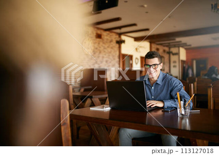 Authentic portrait of young smiling businessman looking at camera with laptop in cafe. Hipster like man in stylish glasses and smart casual shirt doing his work on project. Smart casual wear. 113127018