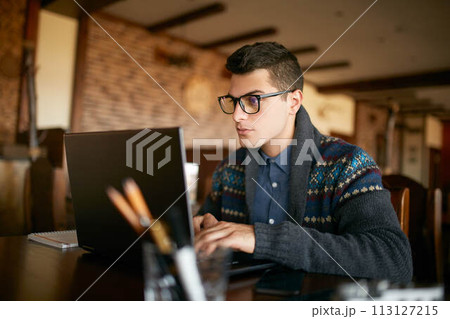 Handsome freelancer businessman in glasses and winter sweater diligently working on laptop in cafe. Man typing on keyboard and searches new job on internet at coffeeshop. Business concept Handsome freelancer businessman in glasses and winter sweater diligently working on laptop in cafe. Man typing on keyboard and searches new job on internet at coffeeshop. Business concept 113127215