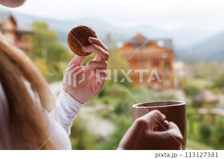 Woman holds cup of coffee and oat cookie in her hands at mountain resort. Female with tea mug and snack with forest and wooden hotel on background. Diet concept. Woman holds cup of coffee and oat cookie in her hands at mountain resort. Female with tea mug and snack with forest and wooden hotel on background. Diet concept. 113127381