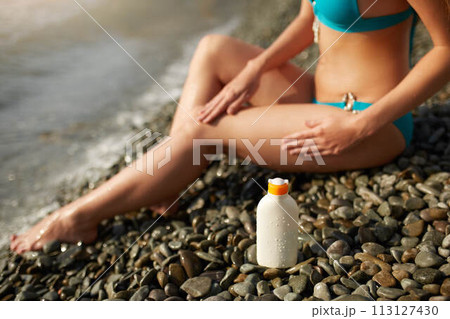 Slim woman tanned lower body in shape lying on pebble beach near sea waves and surf with sunblock cream bottle. Girl applies water resistant sunscreen lotion on legs relaxing in sun holiday. Slim woman tanned lower body in shape lying on pebble beach near sea waves and surf with sunblock cream bottle. Girl applies water resistant sunscreen lotion on legs relaxing in sun holiday. 113127430