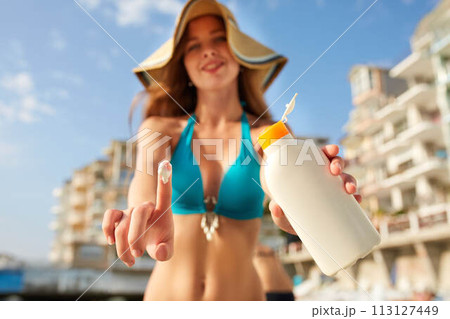 Closeup of smiling caucasian woman showing sunscreen oil bottle and using sun protection cream smear on finger. Pretty tanned girl wearing bikini swimsuit, straw hat and sunglasses on beach near sea. Closeup of smiling caucasian woman showing sunscreen oil bottle and using sun protection cream smear on finger. Pretty tanned girl wearing bikini swimsuit, straw hat and sunglasses on beach near sea. 113127449
