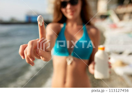 Closeup of smiling caucasian woman showing sunscreen oil bottle and using sun protection cream smear on finger. Pretty tanned girl wearing bikini swimsuit, straw hat and sunglasses on beach near sea. 113127456