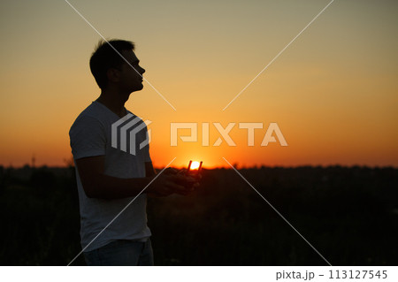 Silhouette of a man piloting drone in the air with a remote controller in his hands on sunset. Pilot takes aerial photos and videos with quadcopter from above Silhouette of a man piloting drone in the air with a remote controller in his hands on sunset. Pilot takes aerial photos and videos with quadcopter from above 113127545