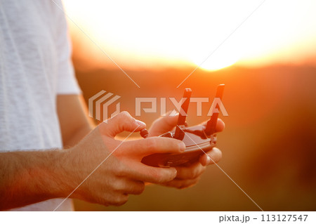 Man holds a drone remote controller in his hands. Close-up of quadrocopter RC during flight. Pilot takes aerial photos and videos with quad on sunset 113127547