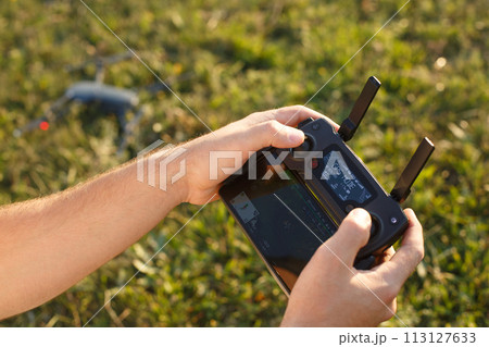 Man holds a drone remote controller in his hands. Quadcopter is on the ground ready to go and fly blurred on the background. Pilot is going to start motors and take off. Man holds a drone remote controller in his hands. Quadcopter is on the ground ready to go and fly blurred on the background. Pilot is going to start motors and take off. 113127633