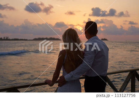 Back view of newly married hug each other and watch sunset skyline on tropical island beach on Maldives. Turquoise ocean lagoon on background. Honeymoon concept. Back view of newly married hug each other and watch sunset skyline on tropical island beach on Maldives. Turquoise ocean lagoon on background. Honeymoon concept. 113127756