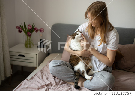 Young blonde 30s woman is resting with a Stripped White cat on the bed at home one spring day. Morning at home. Cute cat licking fingers of female owner in daytime at home. Feeding. 113127757