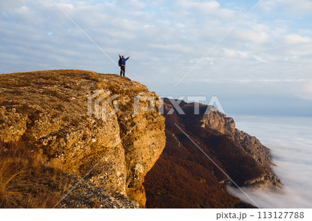 Hiker man cheering with arms spread and raised to the sky after hiking mountain top summit. Climber with backpack on the peak above the clouds. Winner, success, freedom concept. 113127788