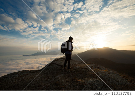 Silhouette of tourist hiker with backpack standing on the top of the mountain and looking at beautiful yellow autumn landscape sunset over clouds 113127792