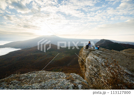 Smiling man hiker with backpack siting and relaxing on the top of the mountain and looking at beautiful yellow autumn landscape sunset over clouds 113127800