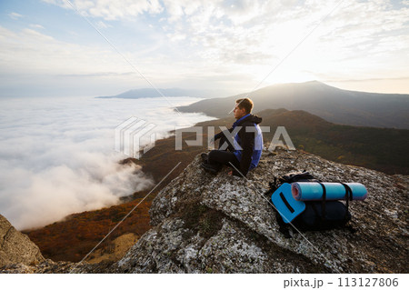 Young tourist hiker with backpack siting and relaxing on the top of the mountain and looking at beautiful yellow autumn landscape sunset over clouds 113127806