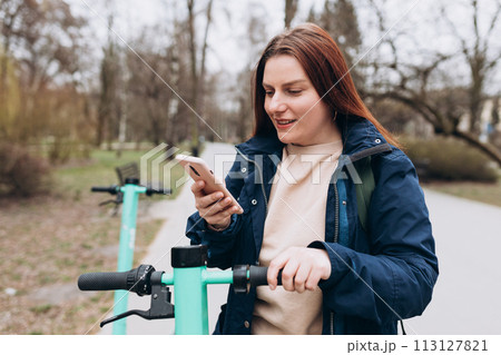 Smiling young woman standing near electric push scooter using smart phone. Ecological transport. Active lifestyle. Urban lifestyle concept. Smiling young woman standing near electric push scooter using smart phone. Ecological transport. Active lifestyle. Urban lifestyle concept. 113127821