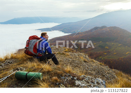 Young tourist hiker with backpack siting and relaxing on the top of the mountain and looking at beautiful yellow autumn landscape sunset over clouds 113127822