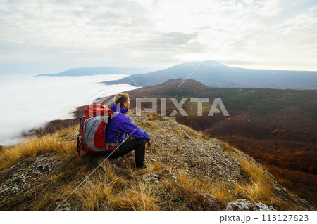 Young tourist hiker with backpack siting and relaxing on the top of the mountain and looking at beautiful yellow autumn landscape sunset over clouds 113127823