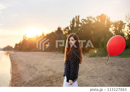 Portrait of young woman with red air balloon walking near the calm sea or lake shore on sunset. Girl on her birthday at the beach. Copyspace. Holiday concept. 113127883