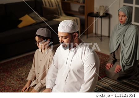 High angle view selective focus shot of modern Muslim man, his wife and son sitting together in living room praying salah 113128051