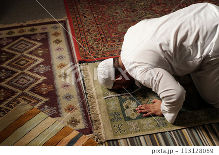 High angle view of mature Muslim man wearing white thobe and tuqiyah doing sajdah on prayer rug during namaz, copy space 113128089