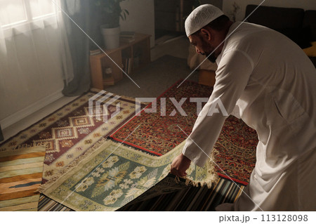 High angle view of Muslim man laying green prayer rug on floor in living room before doing namaz, copy space 113128098
