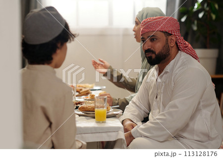 Selective focus shot of Muslim man wearing kandora and keffiyeh sitting festive dining table chatting with his nephew during family dinner 113128176