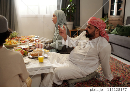 Bearded Muslim man wearing kandora and keffiyeh sitting relaxed at festive table in majilis room talking with family members Bearded Muslim man wearing kandora and keffiyeh sitting relaxed at festive table in majilis room talking with family members 113128177