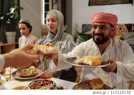 Selective focus shot of cheerful Muslim man sitting at table sharing flatbread during Uraza Bayram celebration dinner 113128178