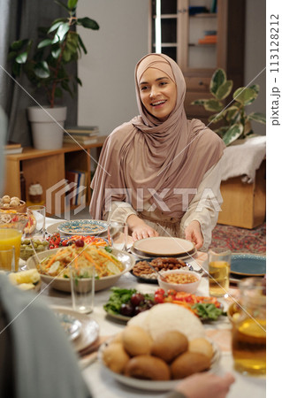 Vertical shot of joyful young Muslim woman wearing hijab sitting at festive table talking to her relatives on Uraza Bayram day Vertical shot of joyful young Muslim woman wearing hijab sitting at festive table talking to her relatives on Uraza Bayram day 113128212