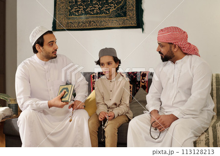 Modern Muslim men and boy sitting on couch in living room discussing suwar from Quran at family gathering on Islamic holiday Modern Muslim men and boy sitting on couch in living room discussing suwar from Quran at family gathering on Islamic holiday 113128213