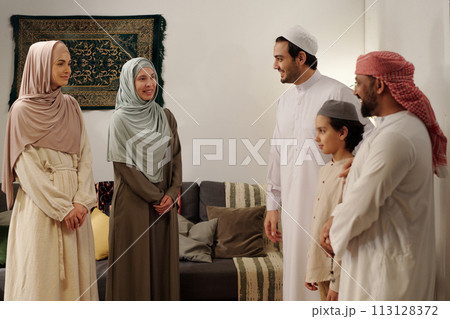 Modern Muslim men, women and kid wearing modest clothes standing in living room having conversation at family gathering on Eid Al-Fitr 113128372