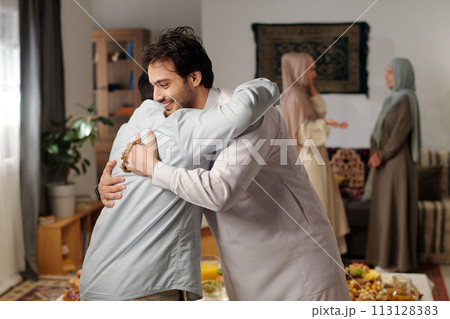 Medium side view of two Muslim men greeting each other with hug on Eid Al-Fitr, two women wearing hijabs talking in background 113128383
