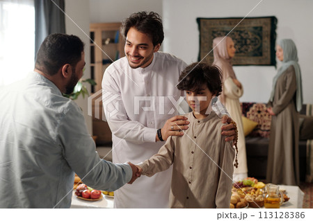 Medium shot of modern Muslim man and his son wearing thobes greeting male relative on Eid al-Fitr, two women talking in background Medium shot of modern Muslim man and his son wearing thobes greeting male relative on Eid al-Fitr, two women talking in background 113128386