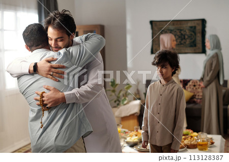 Medium shot of young Muslim man wearing thobe greeting male relative with hug on Eid al-Fitr, his son standing next to him Medium shot of young Muslim man wearing thobe greeting male relative with hug on Eid al-Fitr, his son standing next to him 113128387