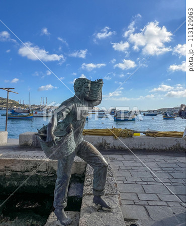 A wonderful monument on the shore of Marsaxlokk, Malta: a young son and daughter meet a father who is carrying two wicker baskets full of caught fish. 113129963