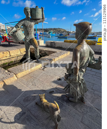 A wonderful monument on the shore of Marsaxlokk, Malta: a young son and daughter meet a father who is carrying two wicker baskets full of caught fish. 113129966