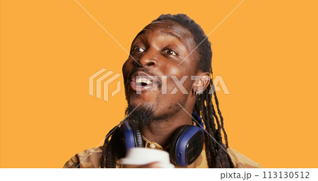 Cheerful guy serving hot cup of coffee in studio, enjoying beverage to gain more energy. African american smiling man drinking caffeine beverage on camera, feeling relaxed and pleased. Cheerful guy serving hot cup of coffee in studio, enjoying beverage to gain more energy. African american smiling man drinking caffeine beverage on camera, feeling relaxed and pleased. 113130512