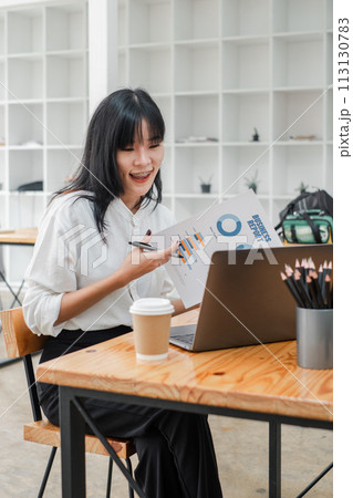 Asian businesswoman with braces excitedly discussing a financial report during a video call in a minimalist office. 113130783