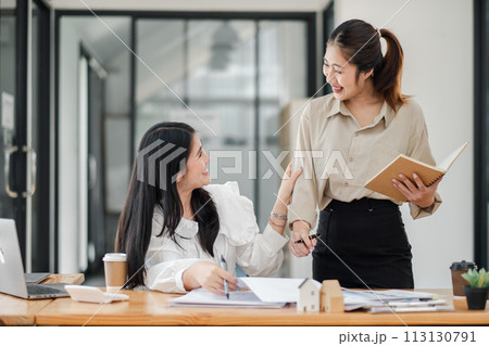 Two engaged female colleagues in a light hearted discussion with notepad and papers at an office desk. 113130791