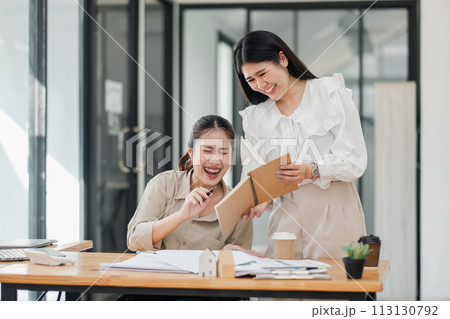 Two engaged female colleagues in a light hearted discussion with notepad and papers at an office desk. 113130792