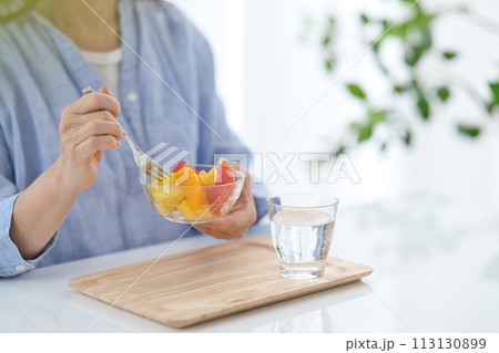 水のグラスを手にフルーツを食べるミドル女性の手元 113130899