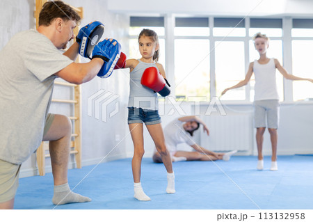 Tween girl in boxing gloves punching pads in hands of father in gym 113132958