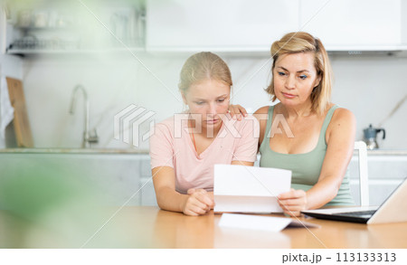 Upset teenage girl reding papers with worried mother in home kitchen 113133313