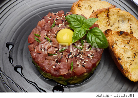 Tuna and avocado tartare with sesame seeds, capers and egg yolk on a dark ceramic plate with baguette bread crouton chips 113138391