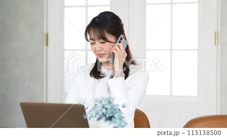 オフィスでノートパソコンを見ながらスマホで通話するミドル女性 113138500