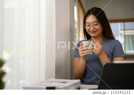 Happy young Asian female freelancer enjoying her cup of coffee while working at home office Happy young Asian female freelancer enjoying her cup of coffee while working at home office 113141131