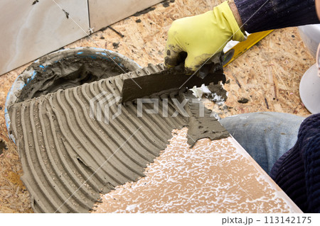 Tiler spreads cement mortar onto tile surface using notched trowel. 113142175