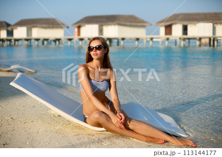 Young woman relaxing in a modern deck chair on a tropical beach. Girl is sitting on a beach sun bed chilling near ocean, palms and bungalows. Vacation concept Young woman relaxing in a modern deck chair on a tropical beach. Girl is sitting on a beach sun bed chilling near ocean, palms and bungalows. Vacation concept 113144177