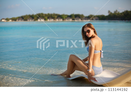 Young woman relaxing in a modern deck chair on a tropical beach with glasses on. Girl is sitting on a beach sun bed chilling near ocean, palms and bungalows. Copy space 113144192