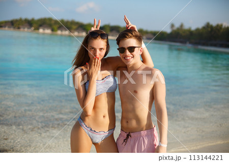 Happy young couple have fun and relax on the beach. Man and woman posing for photo and fool around. Bungalows of spa resort, palms, and blue sea on background. Family travel concept 113144221